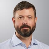 A man with short brown hair and a full beard, wearing a light blue collared shirt, is facing the camera with a neutral expression against a plain, light gray background.