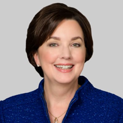 Carrie Penman, smiling, with short brown hair wearing a textured blue jacket against a plain gray background.
