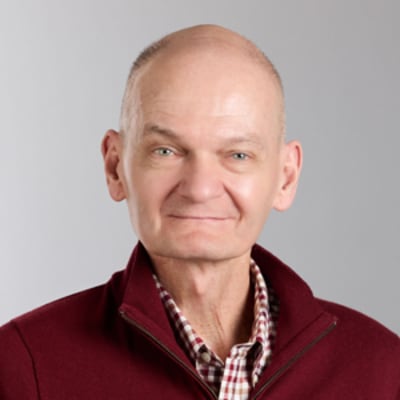 Shon Ramey in a burgundy sweater over a checkered shirt smiling subtly against a light gray background.