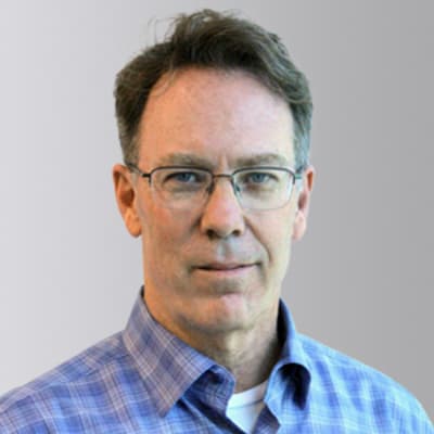 A middle-aged man with short brown hair and glasses, wearing a blue plaid shirt over a white undershirt, stands in front of a neutral gray background.