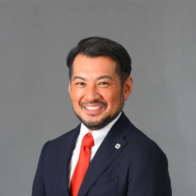 A man in a navy suit with a white shirt and bright red tie stands smiling in front of a plain gray background. He has short black hair and a trimmed beard.