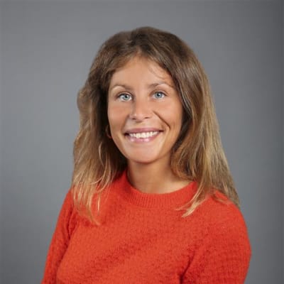 A woman with long, light brown hair, wearing a textured orange sweater, smiles at the camera against a plain gray background.