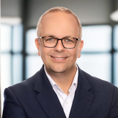 A smiling man wearing glasses and a dark suit jacket stands with arms crossed, leaning against a concrete pillar in a modern, bright office setting with blurred background.