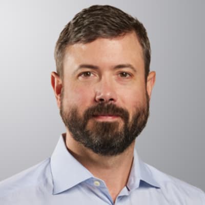 A man with short brown hair and a full beard, wearing a light blue collared shirt, is facing the camera with a neutral expression against a plain, light gray background.