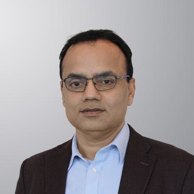 A man with short black hair and glasses wearing a light blue shirt and a dark blazer poses in front of a plain, light gray background.