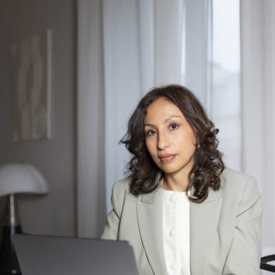 A woman with wavy brown hair, wearing a light gray blazer and white blouse, sits at a table with a laptop in front of her. She looks toward the camera in a modern, well-lit room with sheer curtains.