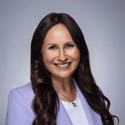 A woman with long brown wavy hair, wearing a lavender blazer with silver buttons and a white shirt, smiles at the camera against a plain gray background.