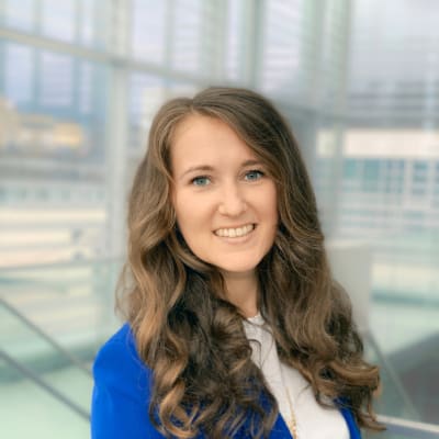 A woman with long brown wavy hair, wearing a lavender blazer with silver buttons and a white shirt, smiles at the camera against a plain gray background.