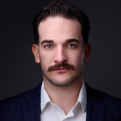 A man with short dark hair and a mustache wearing a white shirt and dark blazer looks directly at the camera against a dark background.