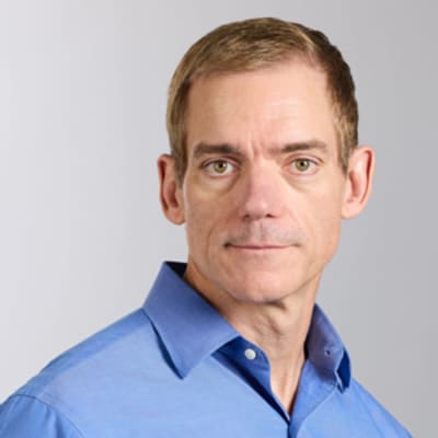 Bob McCarter with short light brown hair in a blue button-up shirt, looking at the camera. The background is plain and light-colored.