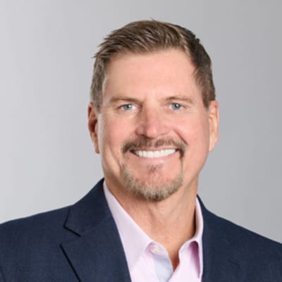 Roger Akers, smiling, with short hair and a beard, wearing a blue blazer and light pink shirt, posed against a light gray background.