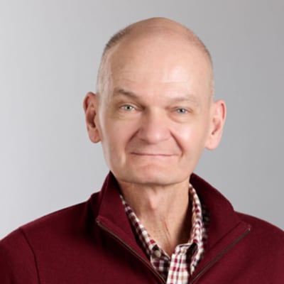 Shon Ramey in a burgundy sweater over a checkered shirt smiling subtly against a light gray background.