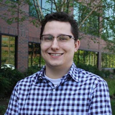 Brian Benston with short brown hair, glasses, and a checkered shirt smiling outdoors in front of a brick building with large windows and green trees.