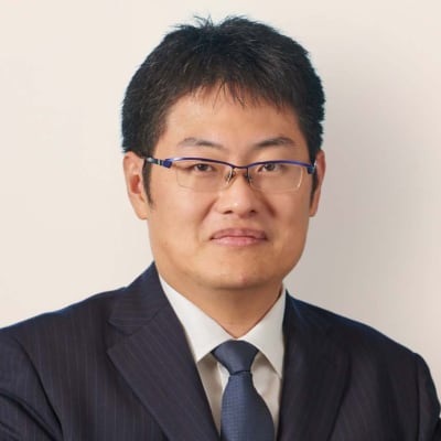 A man wearing glasses and a dark suit with a white shirt and tie, facing the camera and smiling slightly against a plain light background.