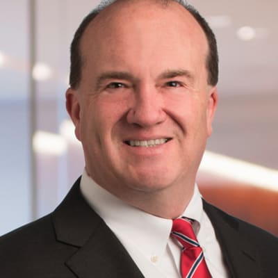 A middle-aged man in a dark suit, white shirt, and red striped tie smiles at the camera in a modern office setting with blurred lights in the background.