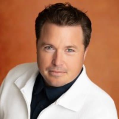 A man with short brown hair wearing a white jacket over a dark shirt poses in front of a warm, orange-brown background. He looks directly at the camera with a slight smile.