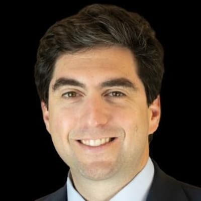 A man with dark hair and a light complexion, wearing a suit and smiling, is pictured against a plain black background.