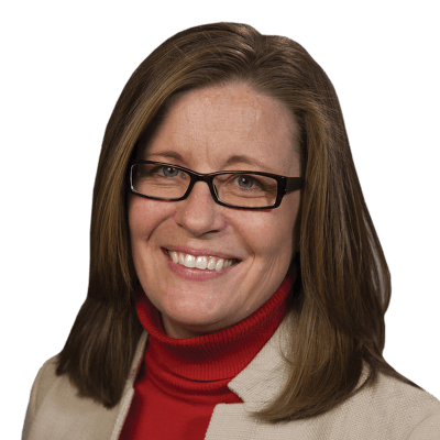 A woman with shoulder-length brown hair wearing glasses, a red turtleneck sweater, and a beige blazer with a navy pocket detail, smiling at the camera.