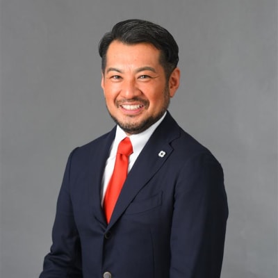 A man in a navy suit with a white shirt and bright red tie stands smiling in front of a plain gray background. He has short black hair and a trimmed beard.