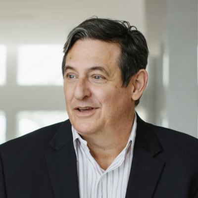 A middle-aged man with dark hair, wearing a dark blazer over a light-colored, striped shirt, is smiling and looking slightly to the side in a bright indoor setting.