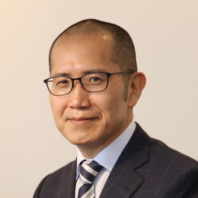 A man in a suit and striped tie, wearing glasses, with short hair and a slight smile, poses against a plain light background.