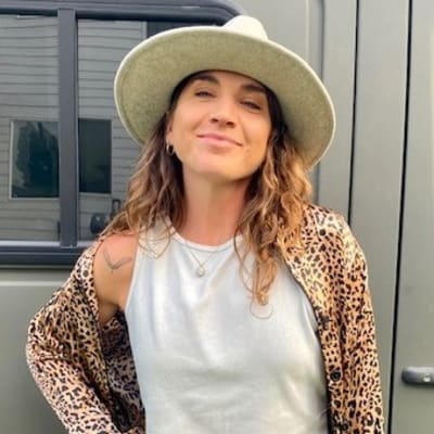 A person with wavy brown hair, wearing a wide-brimmed hat, a sleeveless white top, and a leopard print shirt, stands in front of a green vehicle, smiling confidently.