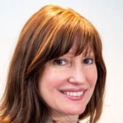 A woman with medium-length brown hair and bangs smiles at the camera. She is wearing a grey jacket over a teal top and is posed against a plain light background.