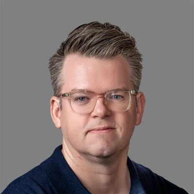 A man with short, neatly styled light brown hair and clear glasses, wearing a dark collared shirt, poses against a plain gray background.