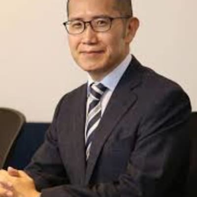 A man wearing glasses and a dark suit with a striped tie sits at a table, smiling slightly, with hands folded in front of him against a neutral background.