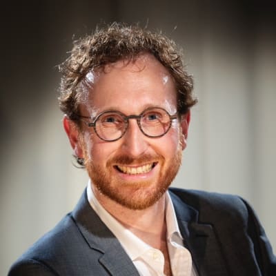 A smiling man with curly brown hair, a short beard, and glasses wearing a dark suit jacket and white shirt, standing in front of a blurred background.