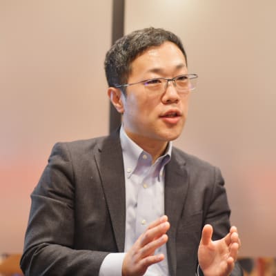 A man wearing glasses and a dark suit gestures with his hands while speaking, sitting in front of a frosted glass background.