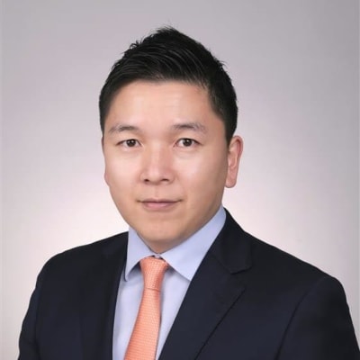 A man in a dark suit, light blue shirt, and orange tie poses for a professional headshot against a plain light gray background.