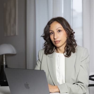 A woman with wavy brown hair, wearing a light gray blazer and white blouse, sits at a table with a laptop in front of her. She looks toward the camera in a modern, well-lit room with sheer curtains.