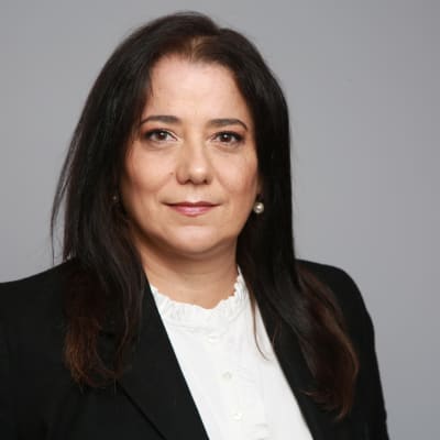 A woman with long dark hair wearing a black blazer and white blouse stands in front of a plain gray background, looking directly at the camera with a neutral expression.