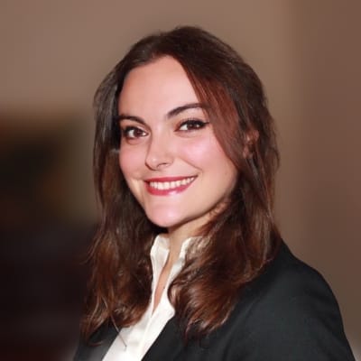 A woman with long brown hair, wearing a black blazer and white shirt, smiles at the camera against a blurred brown background.