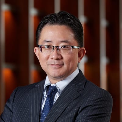 A man in a pinstripe suit, blue tie, and glasses stands with arms crossed, smiling slightly. The background features a wooden panel design with warm lighting.