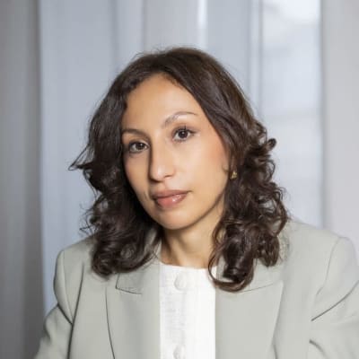 A woman with shoulder-length curly brown hair, wearing a light-colored blazer and white top, sits in front of sheer white curtains and looks at the camera with a neutral expression.