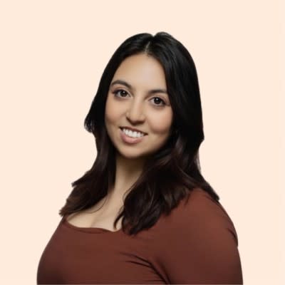 A woman with long dark hair, wearing a brown top, smiles at the camera against a plain light beige background.