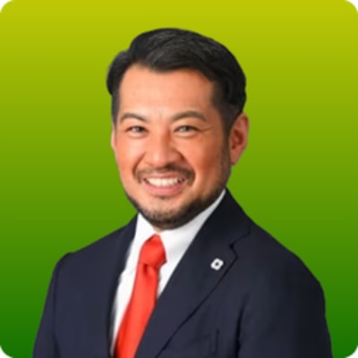 A smiling man with short dark hair and a beard, wearing a navy suit, white shirt, and bright red tie, standing in front of a yellow-to-green gradient background.
