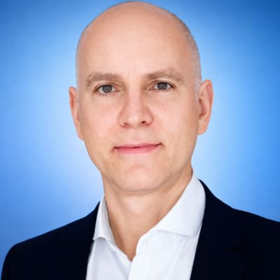A bald man in a dark suit and white shirt poses in front of a smooth blue gradient background, looking directly at the camera with a neutral expression.