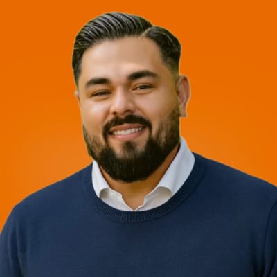 A man with dark hair and a beard, wearing a white collared shirt under a navy sweater, smiles in front of an orange background.