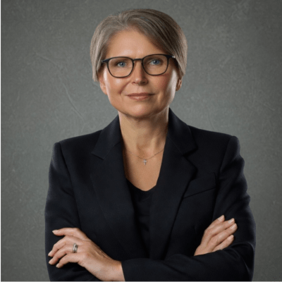 A woman with short gray hair and glasses, wearing a black suit, stands with her arms crossed against a plain gray background.