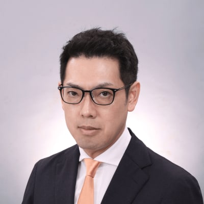 A man with short black hair wearing black glasses, a dark suit, a white shirt, and an orange tie poses against a plain light gray background.