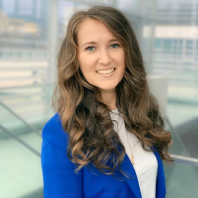 A woman with long brown wavy hair, wearing a lavender blazer with silver buttons and a white shirt, smiles at the camera against a plain gray background.