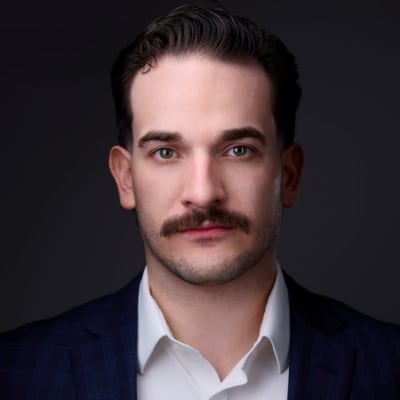 A man with short dark hair and a mustache wearing a white shirt and dark blazer looks directly at the camera against a dark background.