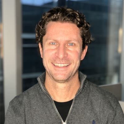 A man with curly brown hair and blue eyes, wearing a dark gray zip-up sweater, smiles at the camera. The background shows large windows and a blurred cityscape.