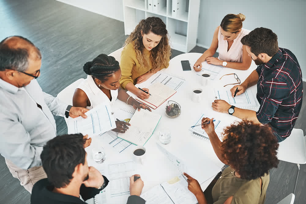 A group of people gathered around a white circular table in an office setting, actively engaged in a meeting.