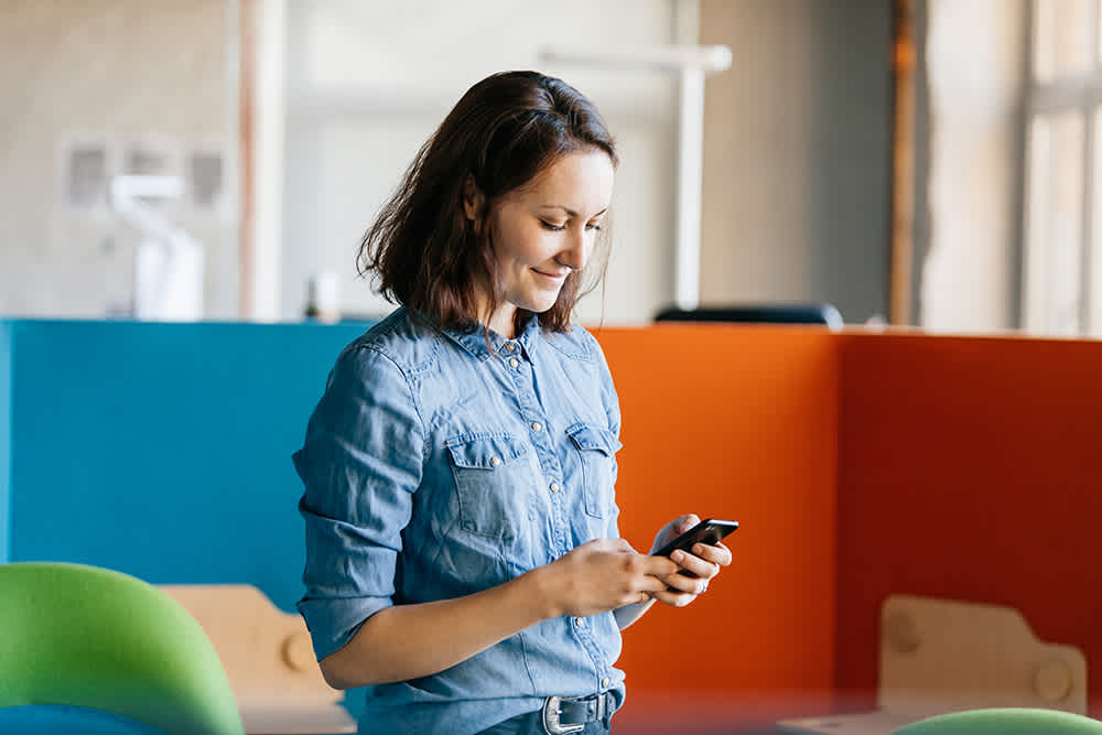 A woman in a denim shirt is looking at her phone and smiling. She is standing in a modern office environment with colorful partitions and natural light streaming in through a window.