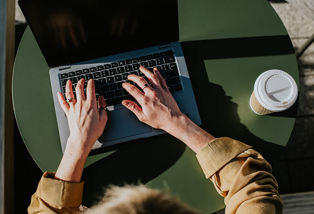Top view of a person typing on a laptop with both hands at a round, green table. A disposable coffee cup with a white lid is placed to the right of the laptop. The person is wearing a brown jacket.