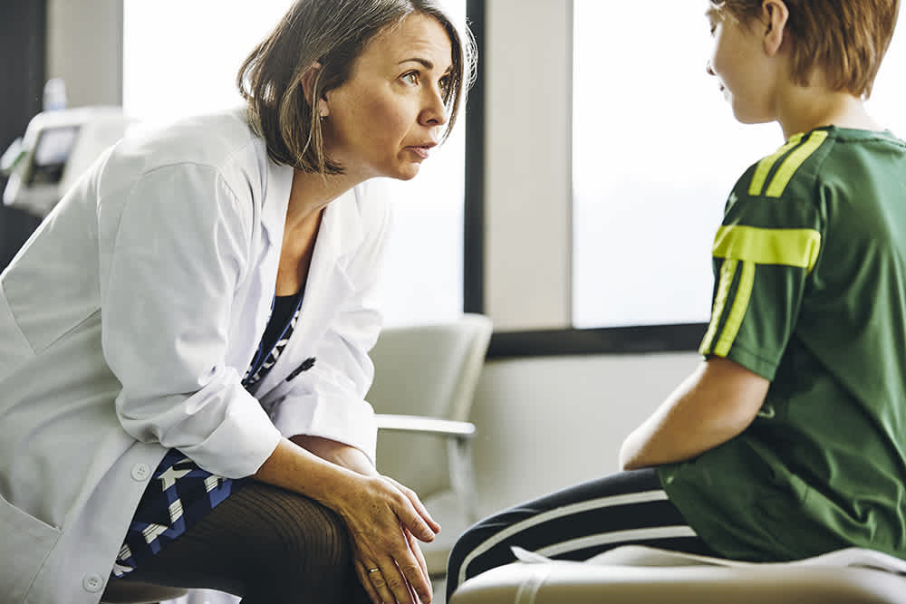 A doctor wearing a white coat is kneeling and attentively listening to a young child sitting on an exam table. The child is dressed in a green and yellow sports jersey with striped pants. The setting is a bright medical office.